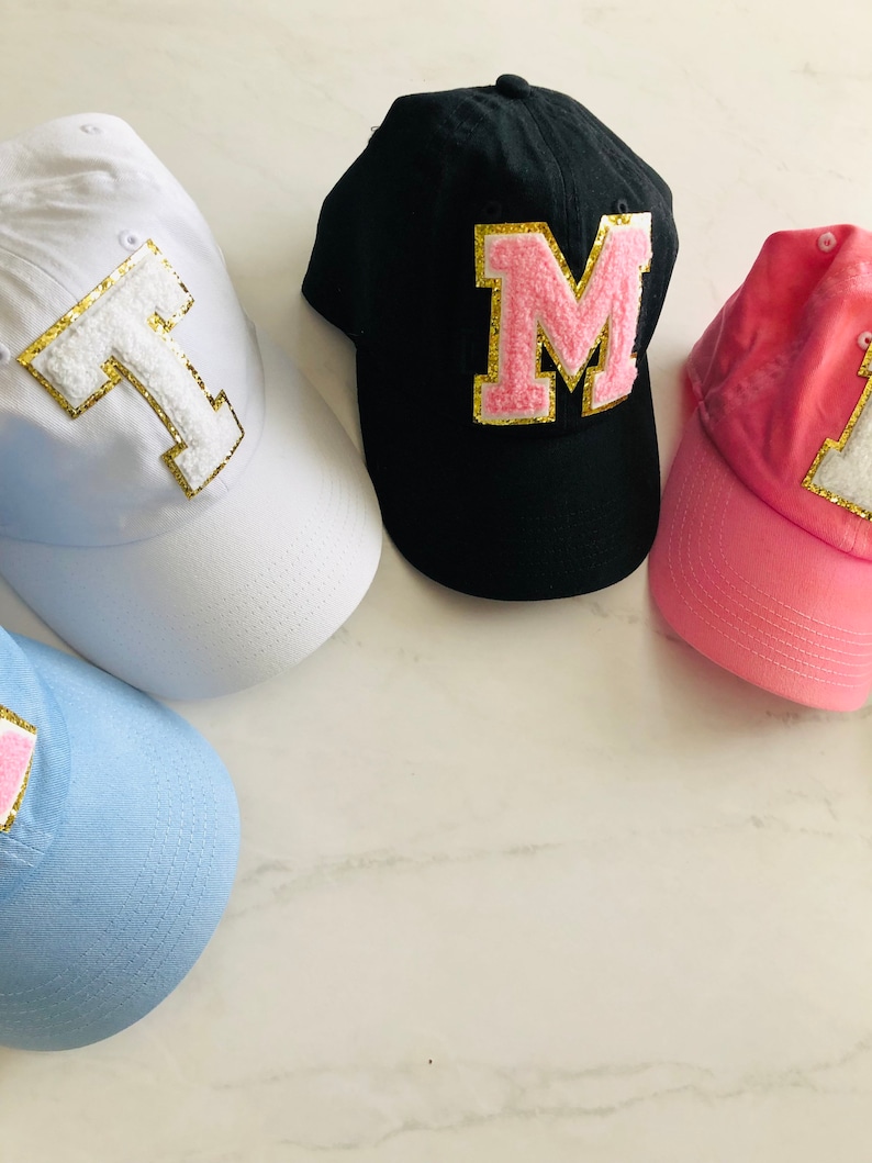Puede incluir: Cuatro gorras de b&eacute;isbol con diferentes colores de tela y grandes letras de fieltro en la parte delantera. Las gorras son blancas, negras, rosas y azul claro. Las letras son T, M, L y J. Las letras est&aacute;n bordeadas con purpurina dorada.