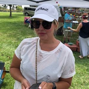 May include: A woman wearing a white t-shirt with distressed detailing, a white baseball cap with the word "Detroit" embroidered on it, and black sunglasses. She is holding a black smartphone in her hand.