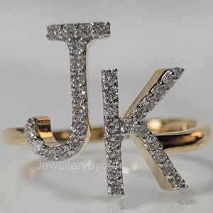 May include: A gold ring featuring the letters "J" and "K" encrusted with small, sparkling diamonds. The ring has a polished gold band and the letters are set with numerous tiny, clear gemstones. The ring is displayed on a reflective white surface.