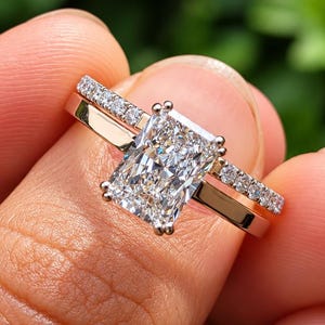 May include: A radiant-cut diamond engagement ring set with a matching pavé band. The central stone is a rectangular, faceted clear diamond held by four prongs on a polished gold band, paired with a thin band featuring small round diamonds.