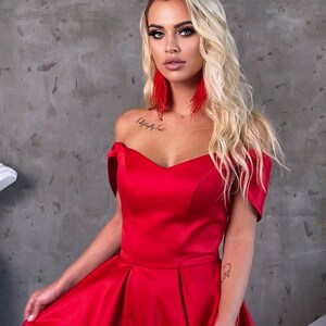 May include: A woman wearing a red off-the-shoulder gown with a sweetheart neckline. The gown has a flowing skirt and is made of a satin-like fabric. The text "Margo Dresses by Margarita Kozak" is visible at the bottom of the image.