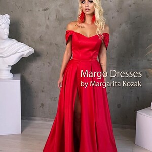 May include: A woman wearing a red off-the-shoulder gown with a thigh-high slit. The gown has a sweetheart neckline and is made of a silky fabric. The text "Margo Dresses by Margarita Kozak" is printed on the dress.