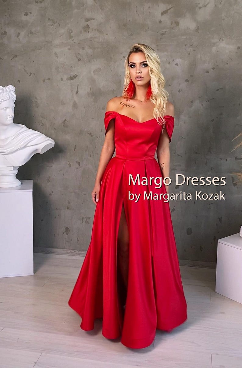 May include: A woman wearing a red off-the-shoulder gown with a thigh-high slit. The gown has a sweetheart neckline and is made of a silky fabric. The text "Margo Dresses by Margarita Kozak" is printed on the dress.