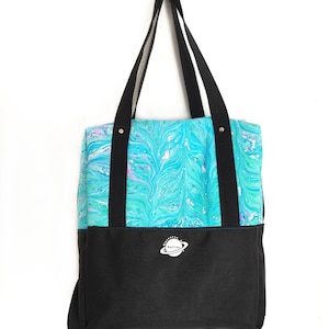 Op de afbeelding: Een draagtas met een bovenkant in een gemarmerd patroon in tinten turquoise, blauw en wit, en een effen zwart onderste gedeelte. De tas heeft zwarte stoffen hengsels en een wit rond logo met een planeet en de tekst ReCreo.