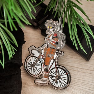 May include: A silver enamel pin featuring a cartoon character riding a bicycle. The character is wearing a helmet, goggles, and a backpack. The bicycle has a white frame and black tires.