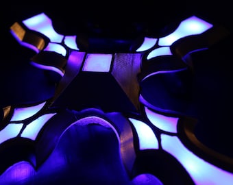 May include: A close-up of a decorative object with a geometric design, illuminated with blue and purple LED lights. The object has a dark, possibly wooden, frame with glowing accents, creating a striking visual effect against a black background.