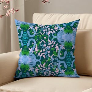 Op de afbeelding: Een vierkant decoratief kussen met een teal achtergrond en een herhalend bloemenpatroon in blauw, groen en roze tinten. Het kussen ligt op een beige bank, met een takje roze bloemen op de achtergrond.