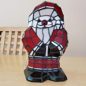 May include: A stained glass Santa Claus lamp with red, white, and black accents. The lamp is made of glass and has a wire cord.
