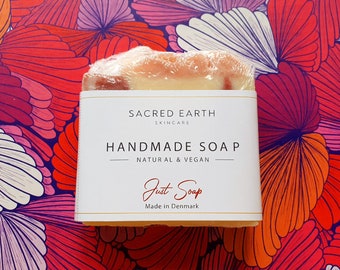 Puede incluir: Una barra de jabón blanca envuelta en plástico con una etiqueta que dice "Sacred Earth Skincare Jabón hecho a mano Natural & Vegano Just Soap Hecho en Dinamarca".