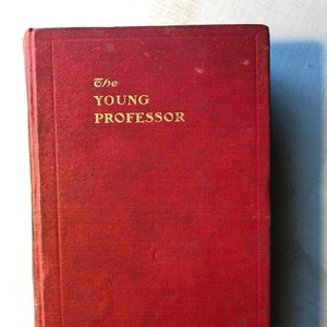 May include: A red hardcover book with the title "The Young Professor" printed in gold lettering on the front cover.