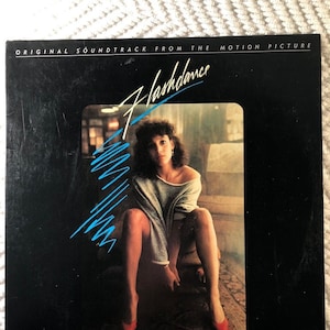 May include: A black vinyl record album cover with the title "Flashdance" in blue and white lettering. The cover features a photo of a woman wearing a gray sweater and red shoes, sitting on a wooden crate.
