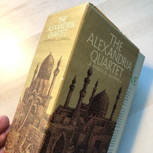May include: A yellow book cover with the title "The Alexandria Quartet" by Lawrence Durrell. The cover features a black and white illustration of a city with minarets and buildings.