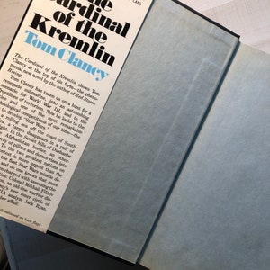 K&ouml;nnte beinhalten: Der Kardinal des Kremls von Tom Clancy, ein Taschenbuch mit einem blauen und wei&szlig;en Umschlag. Der Umschlag zeigt den Titel des Buches in gro&szlig;en, fetten, schwarzen Buchstaben. Der Name des Autors ist ebenfalls in gro&szlig;en, fetten, schwarzen Buchstaben geschrieben. Der Umschlag zeigt auch eine Beschreibung des Buches, die in kleineren, schwarzen Buchstaben geschrieben ist.