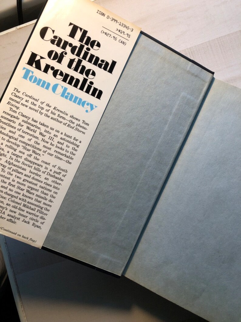 K&ouml;nnte beinhalten: Der Kardinal des Kremls von Tom Clancy, ein Taschenbuch mit einem blauen und wei&szlig;en Umschlag. Der Umschlag zeigt den Titel des Buches in gro&szlig;en, fetten, schwarzen Buchstaben. Der Name des Autors ist ebenfalls in gro&szlig;en, fetten, schwarzen Buchstaben geschrieben. Der Umschlag zeigt auch eine Beschreibung des Buches, die in kleineren, schwarzen Buchstaben geschrieben ist.