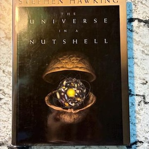 May include: A hardcover book titled The Universe in a Nutshell by Stephen Hawking. The black cover features a gold-toned walnut shell containing a glowing, multi-colored sphere surrounded by thin, intersecting orbital lines in the center.