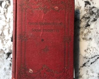 Op de afbeelding: Een vintage rood boek met de titel "Parlor Amusements and Social Etiquette" in gouden letters. De kaft heeft decoratieve goudkleurige versieringen. Het boek vertoont tekenen van slijtage, wat duidt op een antiek item.