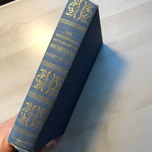 May include: A blue hardcover book with gold lettering on the spine. The title is "The Biographical Dictionary of Musicians" by Rupert Hughes.