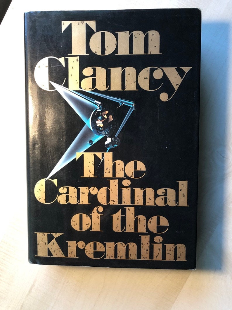 K&ouml;nnte beinhalten: Ein schwarzes Hardcover-Buch mit goldfarbenem Titel "Der Kardinal des Kremls" von Tom Clancy.