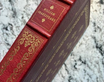 Könnte beinhalten: Ein rotes Hardcover-Buch mit Goldfolienverzierungen auf dem Buchrücken und dem Einband. Der Buchrücken weist erhabene Bünde und den in Gold gedruckten Text "WILLIAM SHAKESPEARE" auf. Filigrane Goldmuster schmücken den Buchrücken und die Kanten.