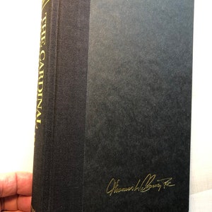 K&ouml;nnte beinhalten: Ein schwarzes Hardcover-Buch mit goldenen Lettern auf dem R&uuml;cken, die "The Cardinal" und "The Kennedy Legacy" lauten. Das Buch ist in goldener Tinte vom Autor "William Colby Jr." signiert.