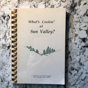 May include: A spiral-bound cookbook titled "What's Cookin' at Sun Valley?" The cover is cream-colored with black text and a green illustration of trees. The text "A collection of favorite recipes from members and friends" is at the bottom.