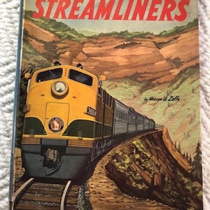 May include: A vintage book cover featuring a yellow and green streamlined train traveling through a mountainous landscape. The book title is "Streamliners" by George H. Ziff.