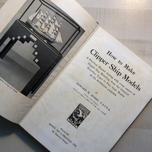 May include: Open book with the title "How to Make Clipper Ship Models." The book features a black and white illustration of a model ship in a display case. The book's pages are aged with a vintage aesthetic.