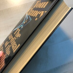 K&ouml;nnte beinhalten: Ein schwarzes Hardcover-Buch mit goldenen Lettern auf dem R&uuml;cken. Der Titel lautet "Der Kardinal des Kremls" von Tom Clancy.