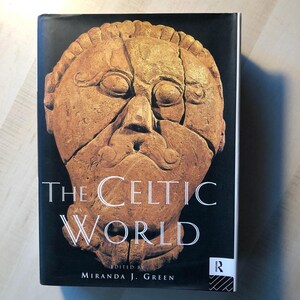May include: The cover of a book titled "The Celtic World" edited by Miranda J. Green. The cover features a close-up of a carved stone face with a prominent nose and a mustache.