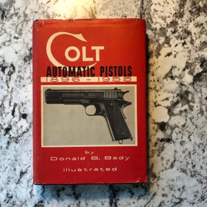 May include: A vintage book titled "COLT Automatic Pistols 1896-1955" by Donald B. Bady. The cover is red with white lettering and a black and white image of a pistol. The book is illustrated.