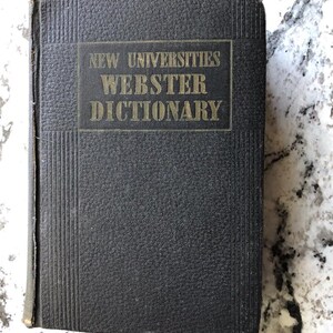 May include: A vintage black Webster's Dictionary with the title "New Universities Webster Dictionary" in gold lettering. The book has a textured cover and is closed. The book is on a white and gray speckled surface.