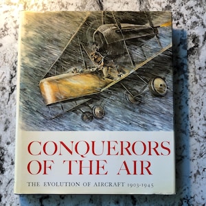 May include: A book cover with the title "CONQUERORS OF THE AIR" in red letters. The cover features an illustration of an early biplane in flight, with a pilot and passenger. The subtitle reads "THE EVOLUTION OF AIRCRAFT 1903-1945".