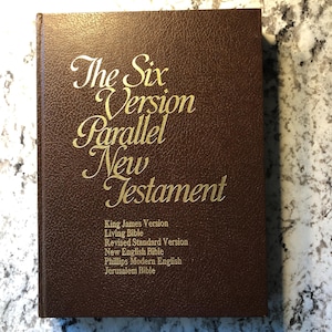May include: A brown hardcover book titled "The Six Version Parallel New Testament" with gold lettering. The book lists six different versions of the New Testament, including the King James Version and the Living Bible.