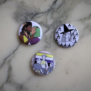 Peut inclure: Trois badges ronds avec des illustrations colorées. L'un représente deux personnes s'embrassant, un autre présente une sorcière avec un chapeau noir, et le troisième montre un groupe de personnes. Les badges sont sur une surface en marbre.