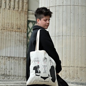 Può includere: Una persona che indossa un cappotto nero e porta una borsa tote bianca con un'illustrazione in bianco e nero di tre donne. La borsa ha il testo "every girl is a riot girl" e "Le Castor Magazine" stampato su di essa.