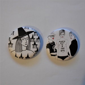 May include: Two round, white buttons with black illustrations. One features a witch with a hat and long hair, surrounded by witch faces. The other depicts three figures with the text "every girl is a riot grrrl".