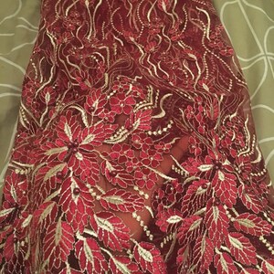 May include: Burgundy lace fabric with gold embroidery and small pearl-like beads. The fabric features a floral design with leaves and flowers.