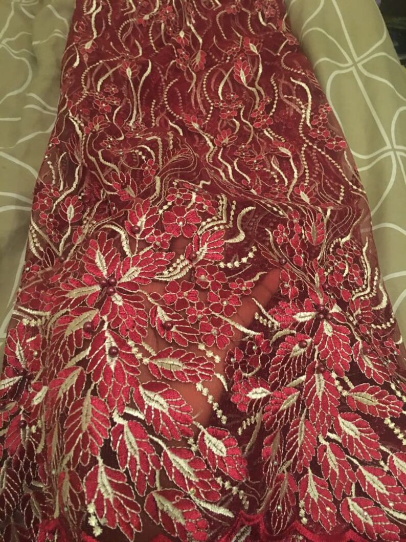 May include: Burgundy lace fabric with gold embroidery and small pearl-like beads. The fabric features a floral design with leaves and flowers.