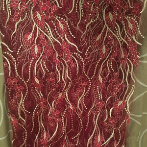 May include: Red lace fabric with gold embroidery featuring a floral pattern. The fabric has a delicate, intricate design with small flowers and leaves.