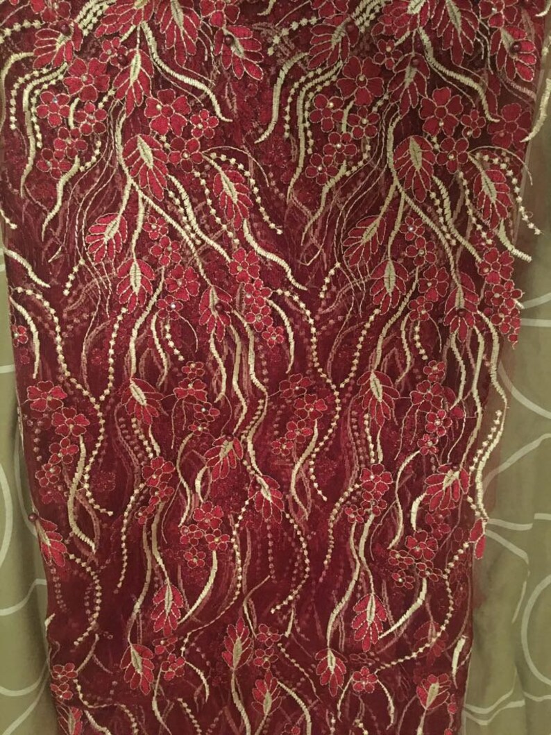 May include: Red lace fabric with gold embroidery featuring a floral pattern. The fabric has a delicate, intricate design with small flowers and leaves.