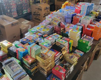 Puede incluir: Una gran variedad de paquetes y cajas de cartas coleccionables, apilados sobre una mesa de madera. Los paquetes son de varios colores, incluyendo rojo, azul, verde y dorado. Se ven cajas de cartón en el fondo.