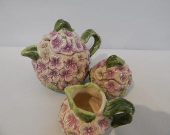Może przedstawiać: Ceramiczny zestaw do herbaty składający się z czajnika z pokrywką, cukiernicy z pokrywką i dzbanuszka na śmietankę. Każdy element jest ozdobiony trójwymiarowymi różowymi i fioletowymi kwiatami hortensji oraz zielonymi liśćmi, wykończony błyszczącym szkliwem.