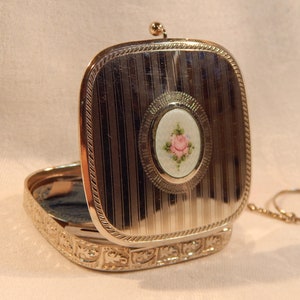 May include: Silver-toned compact with a hinged lid, featuring vertical lines and a decorative border. The lid has an oval inset with a painted pink rose. The open compact reveals a blue interior.