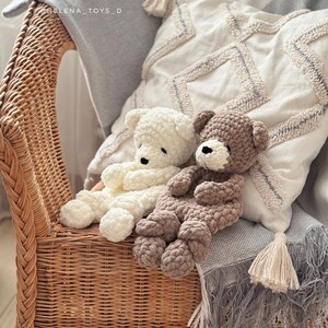 Pode incluir: Dois ursos de pel&uacute;cia de croch&ecirc; feitos &agrave; m&atilde;o, creme e marrom, descansam em uma cadeira de vime. Eles est&atilde;o posicionados em uma almofada decorativa e um cobertor cinza com borlas. Os ursos t&ecirc;m olhos pretos e s&atilde;o feitos de fio macio e texturizado.
