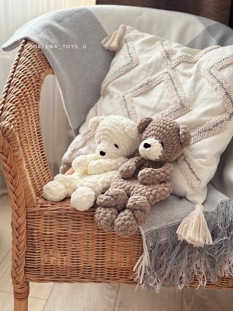 Pode incluir: Dois ursos de pel&uacute;cia de croch&ecirc; feitos &agrave; m&atilde;o, creme e marrom, descansam em uma cadeira de vime. Eles est&atilde;o posicionados em uma almofada decorativa e um cobertor cinza com borlas. Os ursos t&ecirc;m olhos pretos e s&atilde;o feitos de fio macio e texturizado.