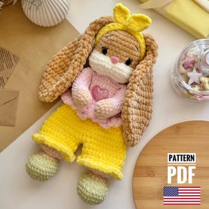 Może przedstawiać: Wzór na szydełku na pluszową zabawkę królika w żółtej opasce, różowej koszulce i żółtych spodniach. Wzór jest dostępny jako plik PDF do pobrania. Obraz zawiera również flagę USA.