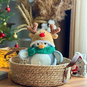 May include: A crocheted penguin wearing a reindeer hat and a green scarf sits in a woven basket with leather handles. The penguin is gray, white, and orange. A small Christmas tree and decorative house are in the background.