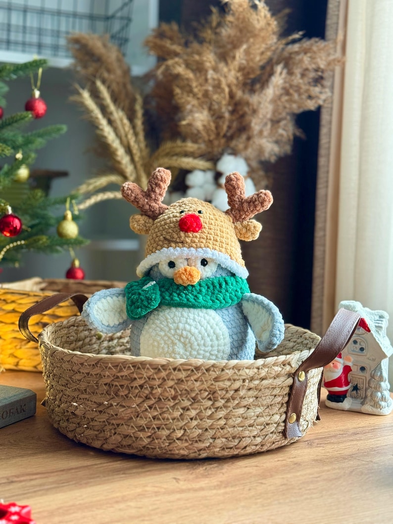 May include: A crocheted penguin wearing a reindeer hat and a green scarf sits in a woven basket with leather handles. The penguin is gray, white, and orange. A small Christmas tree and decorative house are in the background.