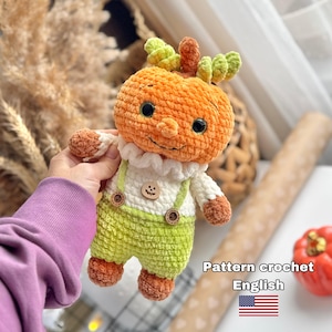 Puede incluir: Una muñeca de ganchillo con cabeza de calabaza naranja, detalles de hojas verdes, cuello blanco con volantes y peto verde claro con botones marrones. La muñeca tiene un pequeño botón de madera con forma de calabaza en el pecho y pies marrones.