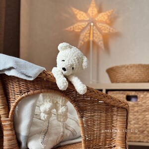 Pode incluir: Um urso de pel&uacute;cia branco feito &agrave; m&atilde;o com olhos pretos est&aacute; sentado em uma cadeira de vime. O urso est&aacute; pendurado na borda da cadeira. Uma lumin&aacute;ria em forma de estrela est&aacute; no fundo.